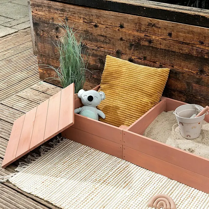Muddy Buddy Sandkasten Space Saver - 150 x 45 x 20 cm, Holz, Puderrosa - Sandkasten aus Holz mit Spielzeug und Kissen. Der Sandkasten ist rosa und steht auf einer Holzterrasse.