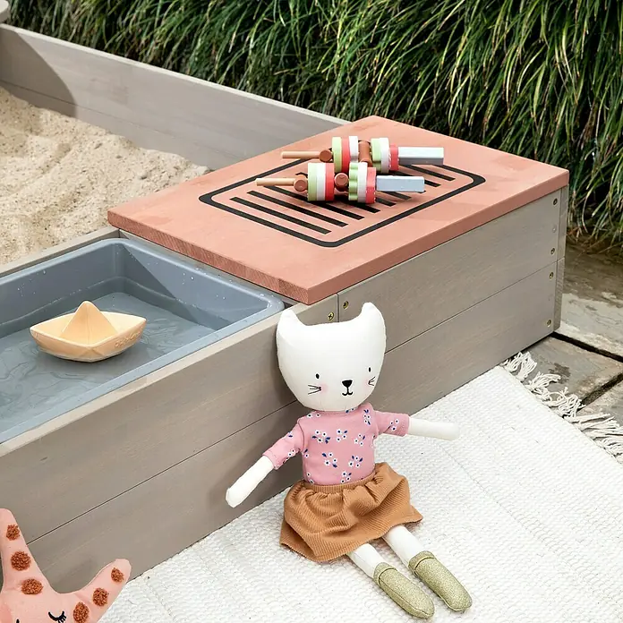 Muddy Buddy Sandkasten Wild Bear - 120 x 120 x 20 cm, Holz, Warmgrau/ Puderrosa - Niedliches Bild von Outdoor-Spielzeug: ein Sandkasten, eine Puppe und Holzspieße auf einem Grill. Ideal für kreatives Spielen im Freien.