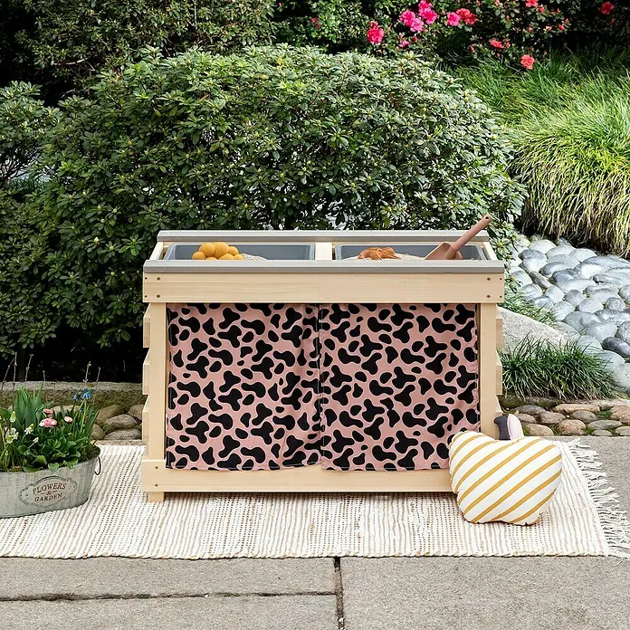 Muddy Buddy Kindertisch zum Matschen Little Leo - L x B x H: 80 x 40 x 55 cm, Holz, Natur/ Puderrosa - Ein hölzerner Spieltisch mit Fächern und niedlichem Kuhmuster-Vorhang, perfekt für den Garten. Ein Herz-Kissen und Blumen verzieren die Szene.