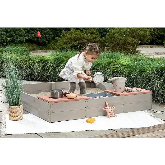 Muddy Buddy Sandkasten Wild Bear - 120 x 120 x 20 cm, Holz, Warmgrau/ Puderrosa - Ein Kind spielt mit dem EXIT Aksent Sand- und Wasserspieltisch mit Spielzeug. Dieser Tisch bietet viel Spaß im Garten!