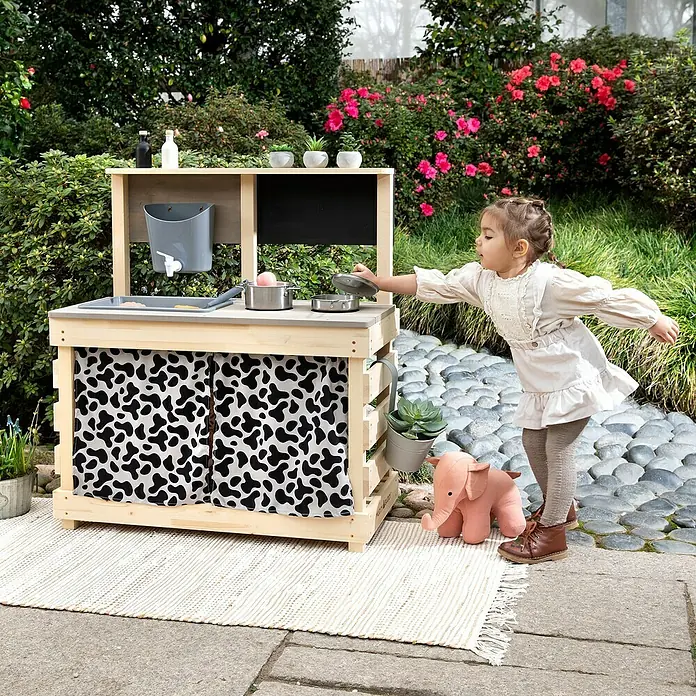 Muddy Buddy Matschküche Big Leo - 80 x 40 x 90 cm, Holz, Natur/ Grau - Ein Kind spielt mit einer Holzküche im Garten, die mit einer Spüle, einem Herd und einer Tafel ausgestattet ist.