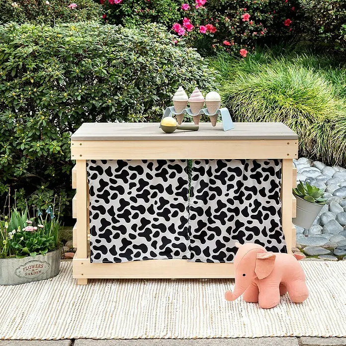 Muddy Buddy Kindertisch zum Matschen Little Leo - L x B x H: 80 x 40 x 55 cm, Holz, Natur/ Grau - Ein Spielstand aus Holz mit einem Kuhmuster-Vorhang, Eiscreme-Requisiten und einem Plüsch-Elefanten vor einem üppigen Garten.
