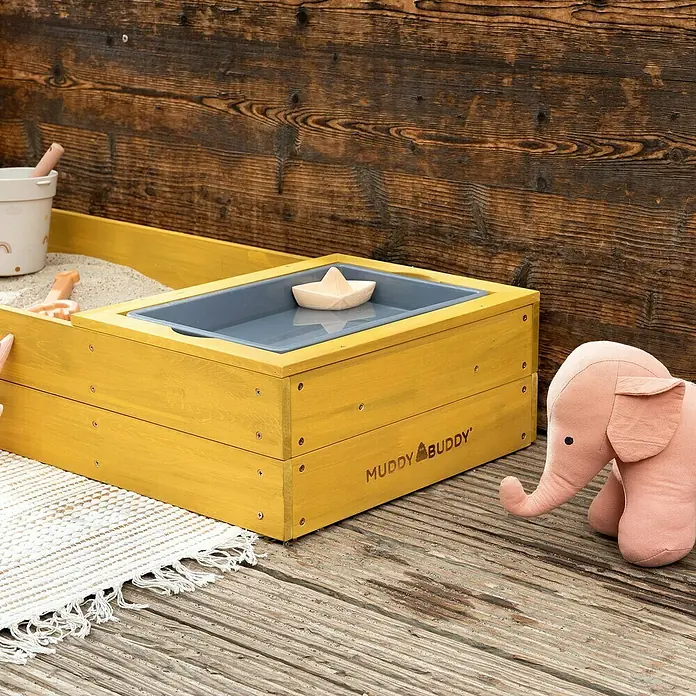 Muddy Buddy Sandkasten Space Saver - 150 x 45 x 20 cm, Holz, Ocker - Eine gelbe Muddy Buddy-Sandkiste mit einer grauen Wasserschale und einem Papierboot darauf, zusammen mit einem rosa Stoffelefanten.
