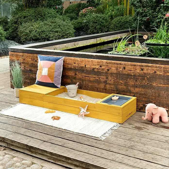 Muddy Buddy Sandkasten Space Saver - 150 x 45 x 20 cm, Holz, Ocker - Ein gelber Holzsandkasten mit Spielzeug steht auf einer Terrasse. Der Sandkasten bietet Platz zum Spielen und Entdecken im Freien.