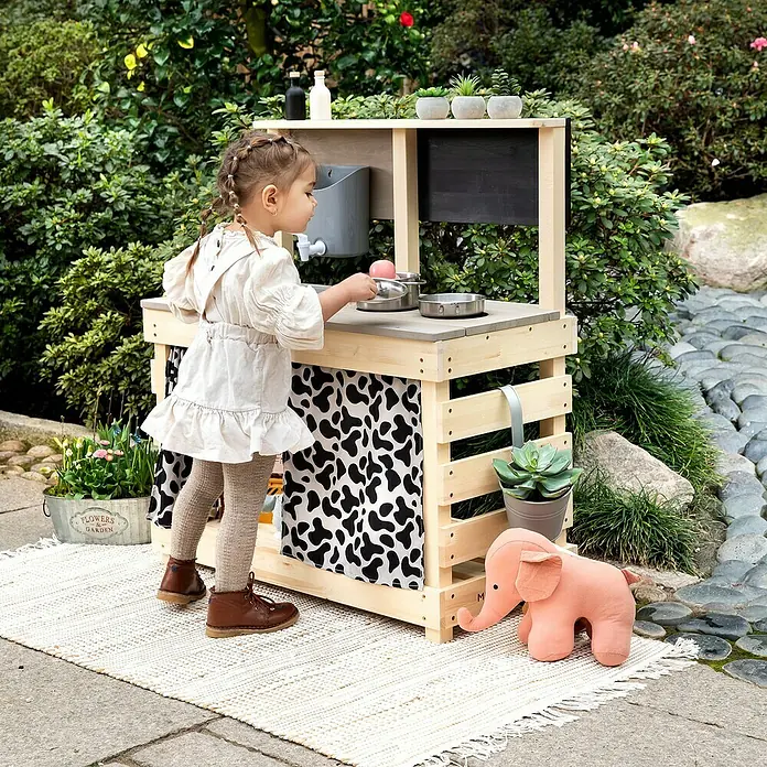 Muddy Buddy Matschküche Big Leo - 80 x 40 x 90 cm, Holz, Natur/ Grau - Ein kleines Mädchen spielt an einer Outdoor-Spielküche aus Holz mit Kochutensilien und Dekoration im Garten.