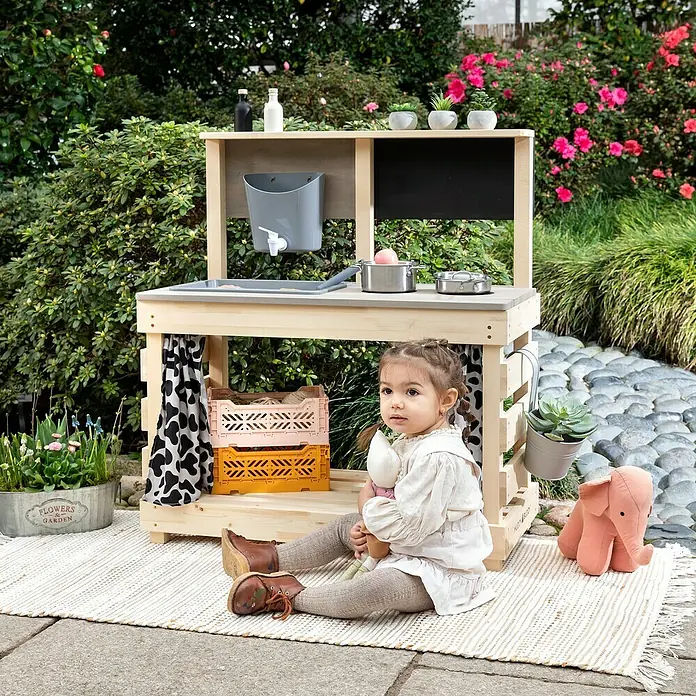 Muddy Buddy Matschküche Big Leo - 80 x 40 x 90 cm, Holz, Natur/ Grau - Ein hölzerner Spielküche für Kinder im Garten, mit Zubehör und einem kleinen Mädchen, das mit einer Puppe spielt.