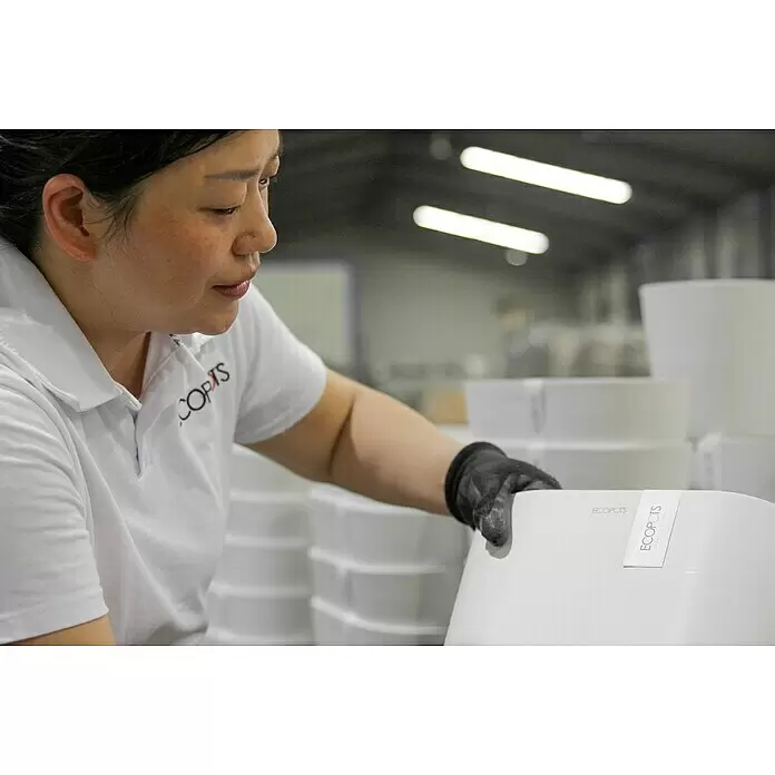 Ecopots Bruges Blumentopf-Untersetzer - Außenmaß (L x B x H): 77,9 x 17,1 x 2,5 cm, Kunststoff, Grau - Weiße ECOPOTS Pflanztöpfe mit Etikett werden von einer Person mit Handschuh in einer Produktionsumgebung gehandhabt.