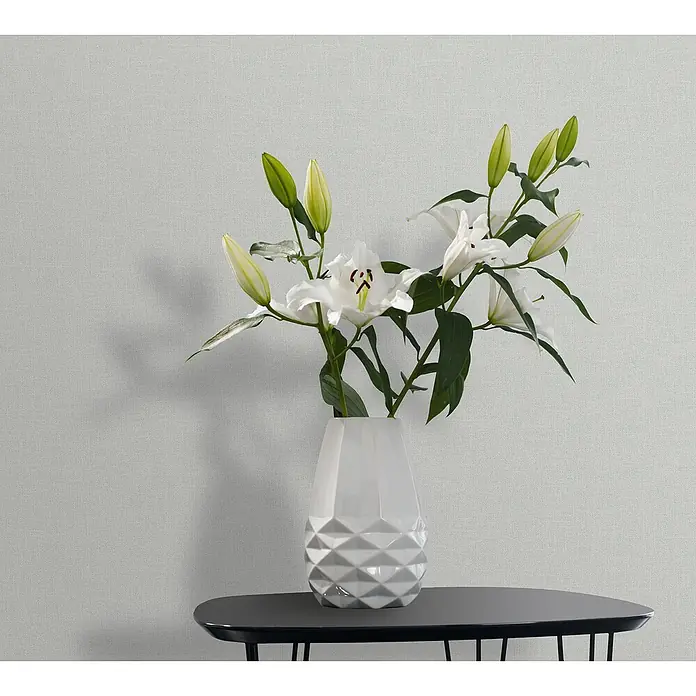 Marburg Heritage Vliestapete Glatte Struktur - Grün, Uni, 10,05 x 0,53 m - Das Bild zeigt eine Nahaufnahme einer weißen Lilienblume in einer weißen Vase, die auf einem Tisch steht. Die Vase hat ein geometrisches Design.
