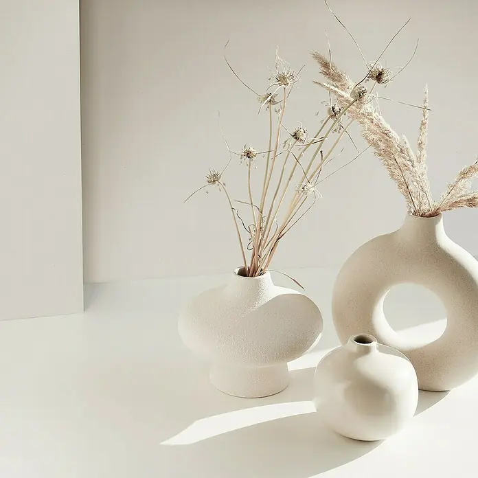 Marburg Struktura Vliestapete Jackie - Beigegrau, Uni, 10,05 x 0,53 m - Elegante Vasen in Weiß, darunter eine Donut-Vase und eine Kugelvase, perfekt für minimalistische Dekorationen mit Trockenblumen.