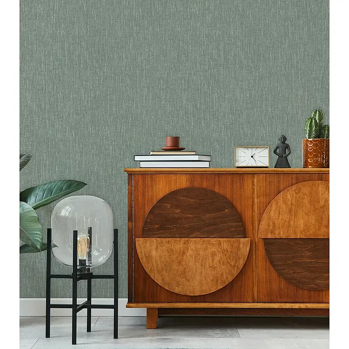 Marburg Heritage Vliestapete Textil-Optik - Grün, Uni, 10,05 x 0,53 m - Einrichtungsszene mit einem Sideboard aus Holz mit einer Uhr, Büchern und einer Pflanze darauf. Im Vordergrund steht eine Stehlampe.