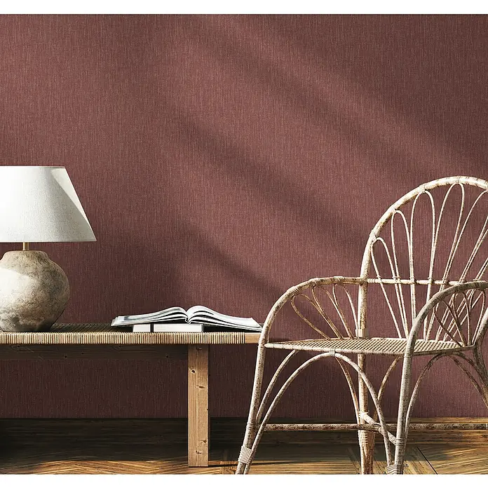 Marburg Heritage Vliestapete Textil-Optik - Rot, Uni, 10,05 x 0,53 m - Einladende Szene mit einer Lampe, einem Buch und einem Korbstuhl vor einer strukturierten Wand.