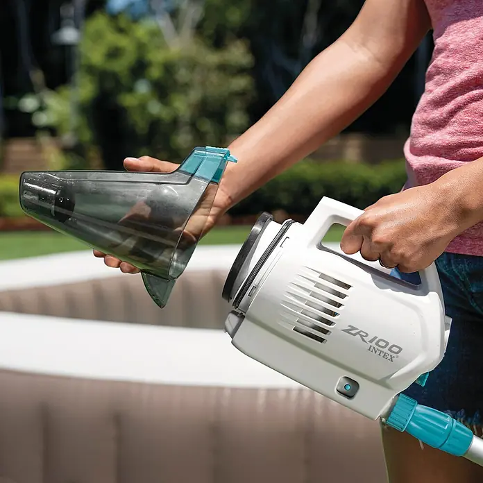 Intex Aspirador para piscina Basic ZR100 - A batería, Específico para: Piscinas elevadas de hasta 5,49 m de diámetro - Aquí hay una aspiradora de piscina Intex ZR100 que está siendo sostenida e inspeccionada por una persona, cuenta con un recipiente de desechos transparente.