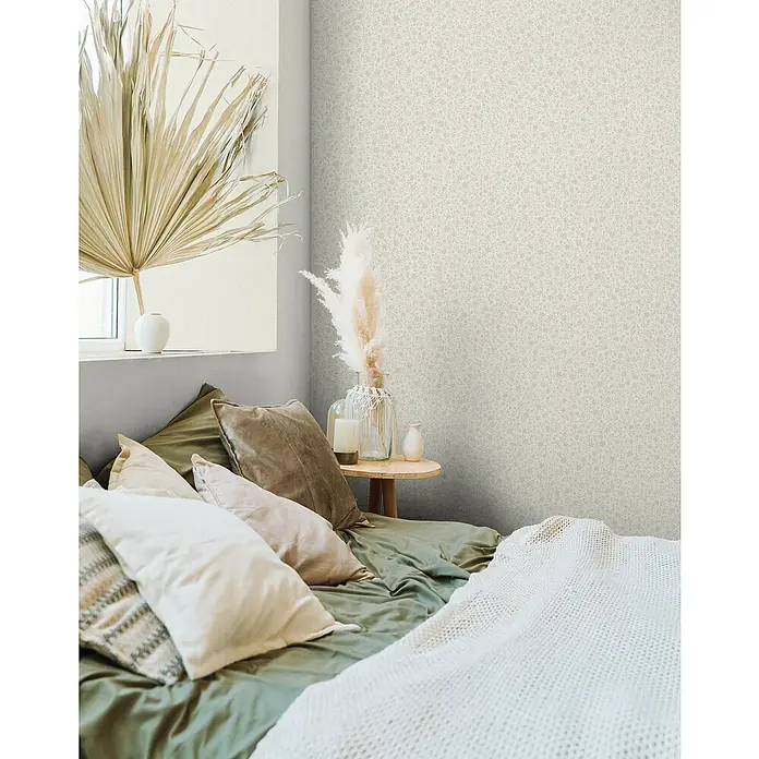 Marburg Heritage Vliestapete mit feinen Blättern - Beige, Floral, 10,05 x 0,53 m - Nahaufnahme eines Schlafzimmers mit einem bequemen Bett, dekoriert mit Zierkissen und einer warmen Decke, vor einer dezent gemusterten Wand.