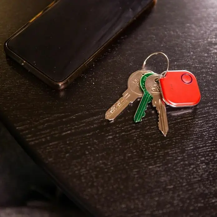 Shelly Schalter BLU Button1 - Rot, Batteriebetrieben - Schlüsselanhänger mit rotem Tile-Tracker und mehreren Schlüsseln auf einem dunklen Tisch neben einem Smartphone.
