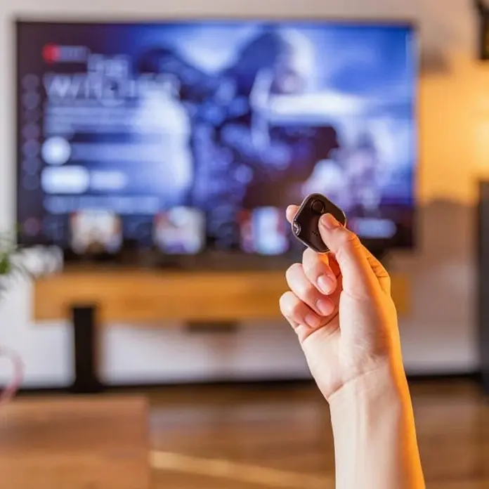 Shelly Schalter BLU Button1 - Schwarz, Batteriebetrieben - Eine Hand hält eine kleine, schwarze Fernbedienung vor einem Fernseher, der "The Witcher" zeigt. Praktisches Zubehör für Mediensteuerung.