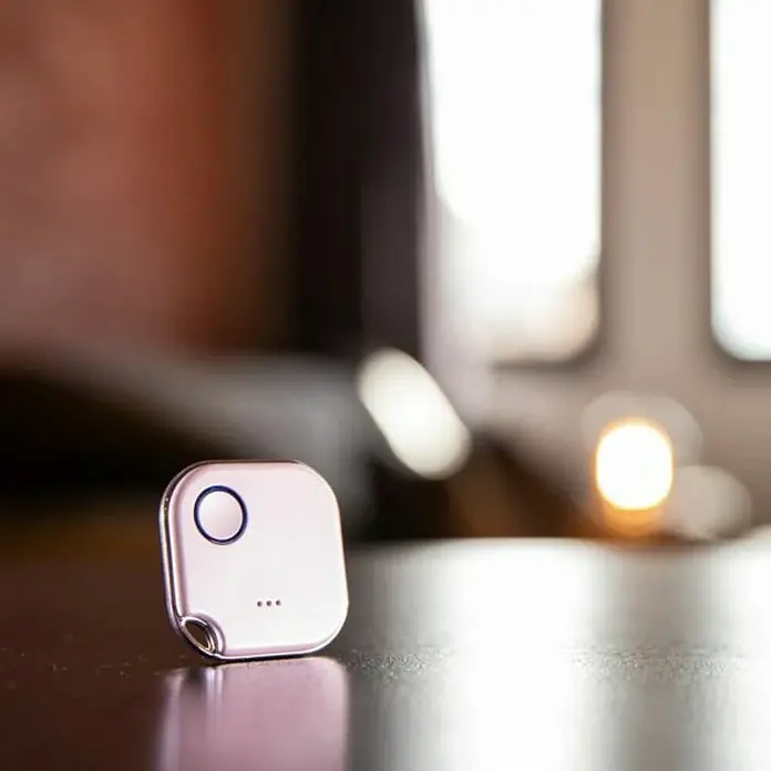 Shelly Schalter BLU Button1 - Weiß, Batteriebetrieben - Nahaufnahme eines quadratischen, rosa-silbernen Schlüsselfinders mit einem Knopf und drei Punkten, der auf einer glänzenden Oberfläche steht.