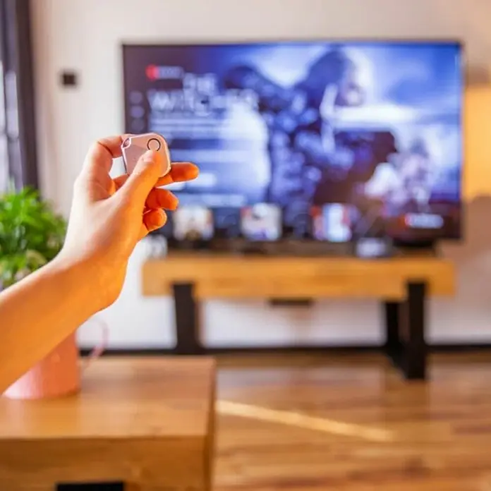 Shelly Schalter BLU Button1 - Weiß, Batteriebetrieben - Nahaufnahme einer Hand, die ein quadratisches, weißes Gadget hält, vor einem verschwommenen Fernseher, das ein modernes, technisches Ambiente vermittelt.