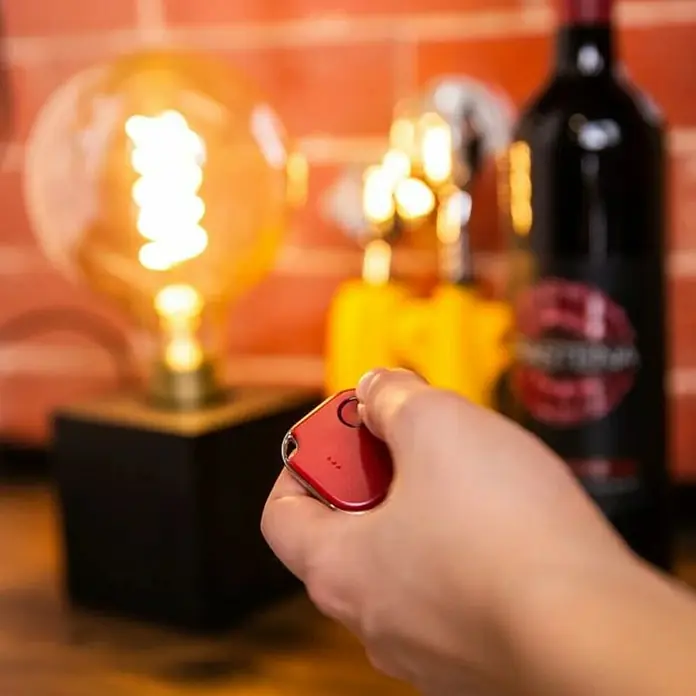 Shelly Schalter BLU Button1 - Rot, Batteriebetrieben - Nahaufnahme eines roten Schlüsselfinders in der Hand, im Hintergrund Lampe, Weinflasche und Backsteinwand.