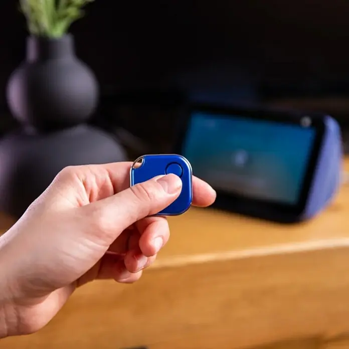 Shelly Schalter BLU Button1 - Blau, Batteriebetrieben - Nahaufnahme eines blauen Tile Mate Trackers in einer Hand, mit einem Smart Home Gerät im Hintergrund.