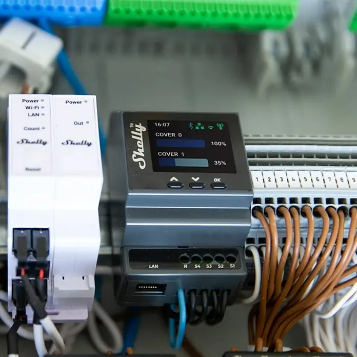 Shelly Schaltcontroller Pro Dual Cover / Shutter PM - 110 V - 240 V, 16 A - Nahaufnahme eines Shelly-Automatisierungsgeräts mit Display, das den Status von Abdeckungen anzeigt, umgeben von Verkabelung und elektronischen Komponenten.