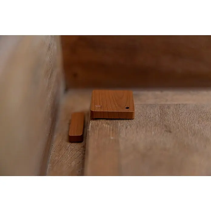 Shelly Fenster- & Türsensor BLU Door/Window - Bluetooth, Braun, Batteriebetrieben - Nahaufnahme eines quadratischen Holzproduktes mit Loch und rechteckigem Accessoire. Minimalistisches Design auf hölzernem Untergrund.