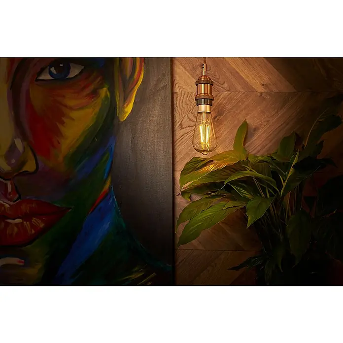 Shelly Smart-LED-Lampe Vintage ST64 - E27, Dimmbar, Warmweiß, 750 lm, 7 W - Ein stimmungsvolles Bild mit einer Edison-Glühbirne, die warmes Licht verbreitet, neben einem farbenfrohen Gemälde und einer grünen Pflanze.