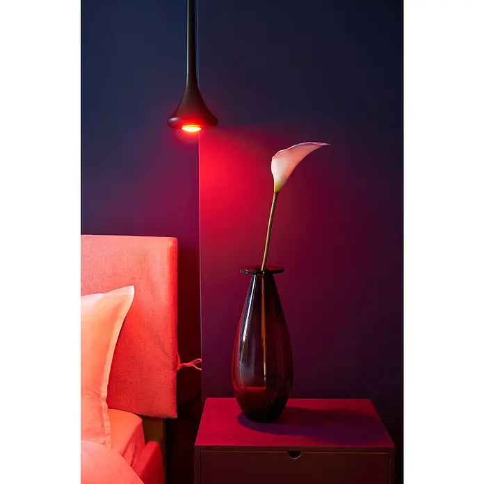 Shelly Smart-LED-Lampe Duo - RGBW GU10 - GU10, Dimmbar, Warmweiß, 400 lm, 5 W - Elegante Glasvase mit einer Calla-Lilie unter einem roten Licht, das eine gemütliche Atmosphäre schafft. Das Nachttischchen ergänzt das moderne Ambiente.