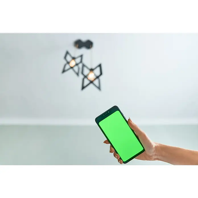 Shelly Smart-LED-Lampe Vintage ST64 - E27, Dimmbar, Warmweiß, 750 lm, 7 W - Ein Smartphone mit grünem Bildschirm wird vor einer modernen Zimmerlampe gehalten. Ideal für Smart Home oder Technologie-Konzepte.