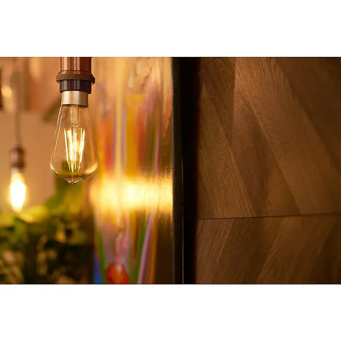 Shelly Smart-LED-Lampe Vintage ST64 - E27, Dimmbar, Warmweiß, 750 lm, 7 W - Nahaufnahme einer dekorativen Edison-Glühbirne, die ein warmes Licht ausstrahlt, mit einem Hintergrund aus Kunst und Holzvertäfelung.