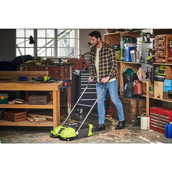 Ryobi ONE+ Akku-Handkehrmaschine R18SW3-0 - 18 V, Ohne Akku, Behältervolumen: 17 l - Ein Mann benutzt einen gelb-schwarzen Kehrbesen, um eine Werkstatt aufzuräumen. Er trägt ein gelb kariertes Hemd, Jeans und Stiefel.