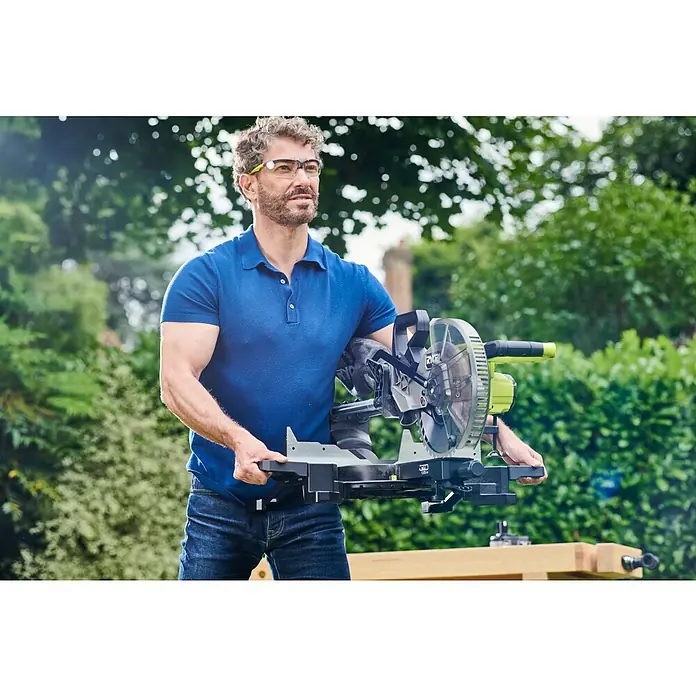 Ryobi ONE+HP Akku-Kapp- & Gehrungssäge RMS25418X-0 - 18 V, Ohne Akku, 4.100 U/min - Ein Mann mit Schutzbrille trägt eine RYOBI Kapp- und Gehrungssäge vor dem Hintergrund eines üppigen Gartens. Die Säge ist das Hauptaugenmerk.