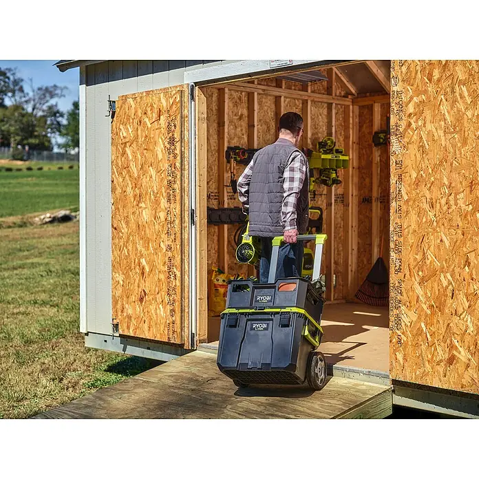 Ryobi Link Werkzeugkoffer RSL201 Trolley - L x B x H: 57 x 49,5 x 39 cm, Kunststoff, 2 Räder - Ein Mann zieht einen schwarzen und grünen Ryobi Werkzeugwagen aus einem Schuppen. Der Wagen hat mehrere Fächer und Räder für einfachen Transport.