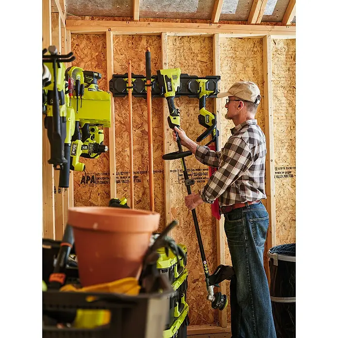 Ryobi Link Wandhalterung RSLW803 - Geeignet für: Handwerkzeuge, Größe: M, Passend für: Ryobi ONE+ Sortiment - Ein Mann nimmt einen RYOBI Rasentrimmer von einer Wandhalterung in einer Garage. Werkzeuge und Gartengeräte sind ordentlich verstaut.