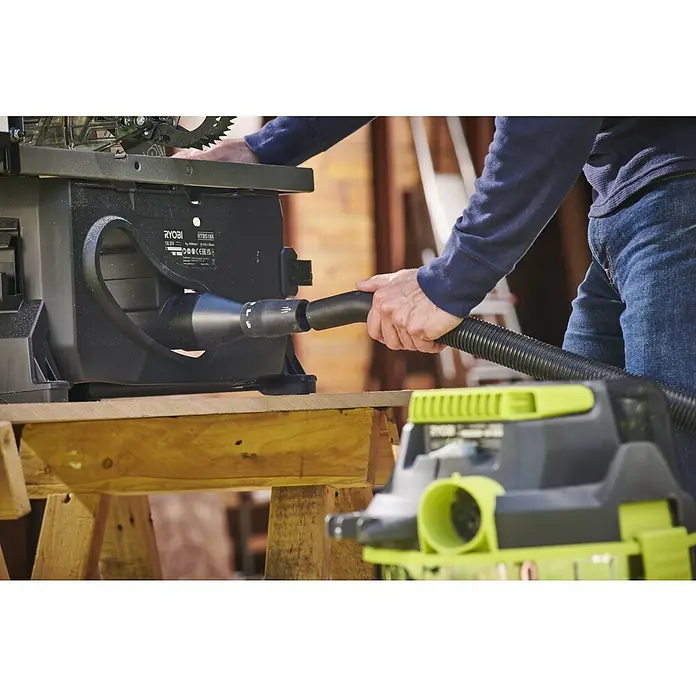 Ryobi ONE+HP Akku-Tischkreissäge RTBS18X-0 - 18 V, Ohne Akku, Durchmesser Sägeblatt: 210 mm - Detailaufnahme einer Ryobi Tischkreissäge mit angeschlossenem Staubsauger, der Staub absaugt, während der Bediener arbeitet.