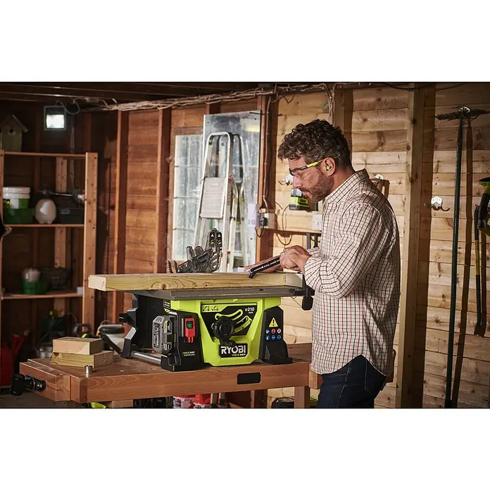 Ryobi ONE+HP Akku-Tischkreissäge RTBS18X-0 - 18 V, Ohne Akku, Durchmesser Sägeblatt: 210 mm - Ein Mann bedient eine Ryobi Tischkreissäge in einer Werkstatt, ausgestattet mit Sicherheitsbrille und Gehörschutz, um ein Holzbrett zu bearbeiten.