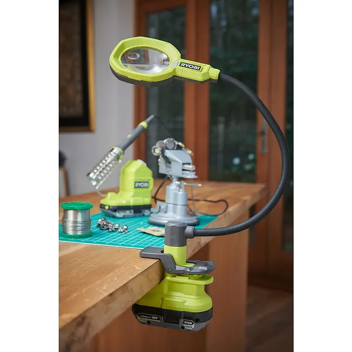 Ryobi ONE+ Akku-Lupenleuchte RML18-0 - 18 V, Ohne Akku - Nahaufnahme einer Ryobi-Arbeitsplatzleuchte mit Schwanenhals, die an einer Werkbank befestigt ist, mit Werkzeugen im Hintergrund.