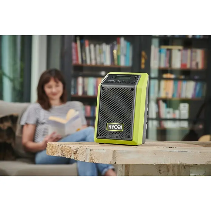 Ryobi ONE+ Akku-Radio RR18-0 Bluetooth® - Ohne Akku - Nahaufnahme eines Ryobi ONE+ Radios auf einem Holztisch mit einer Frau im Hintergrund, die ein Buch liest.