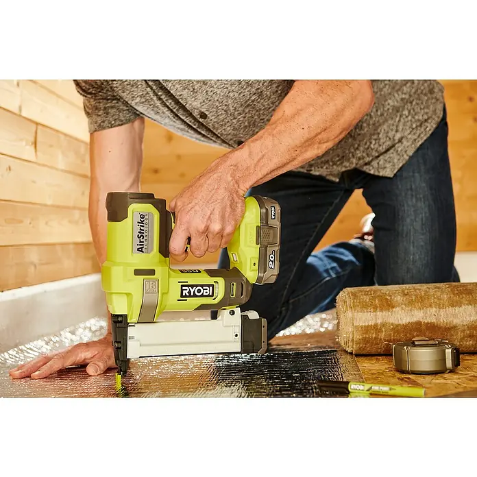 Ryobi ONE+ Maschinenpaket - Akku-Tacker R18GS18-0 + gratis Starter-Kit - Ein Handwerker kniet und benutzt einen gelb-schwarzen Ryobi Airstrike Akku-Nagler für ein Bauprojekt.