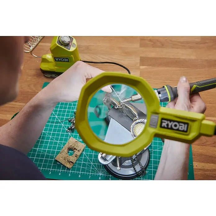 Ryobi ONE+ Akku-Lupenleuchte RML18-0 - 18 V, Ohne Akku - Nahaufnahme einer Person, die mit einem RYOBI Lötkolben an Schmuck arbeitet, durch eine Lupenbrille betrachtet.