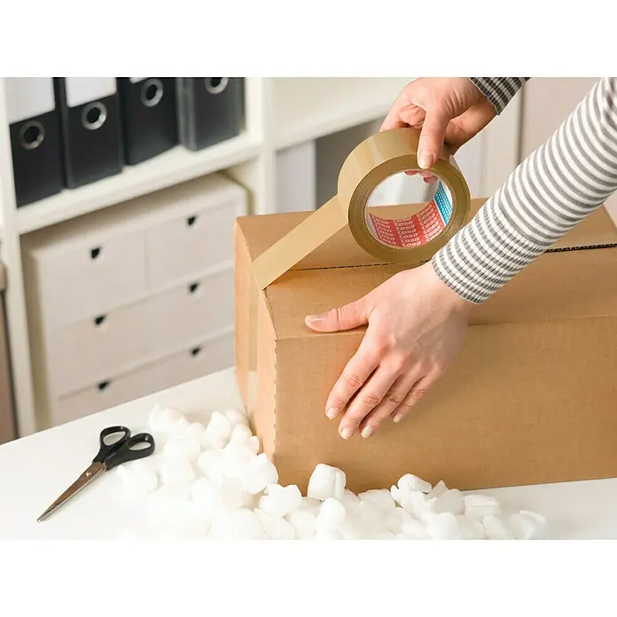 Tesa Cinta de embalaje Strong - 50 mm x 66 m, Marrón - Una persona sella una caja de cartón con cinta de embalar marrón, asegurándola para su envío.