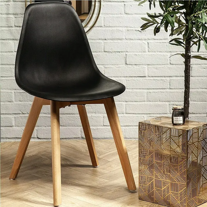 Home Deco Factory Silla Stockholm - Negro, 4 pzs. - Silla de plástico negro con patas de madera junto a una mesa auxiliar dorada y una planta frente a una pared de ladrillo blanco.