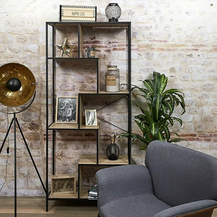 Home Deco Factory Estantería de madera Edison - L x An x Al: 30 x 80 x 180 cm, Negro - Una estantería con artículos decorativos, incluyendo marcos de fotos y una lámpara, contra una pared de ladrillo, con un sillón gris en primer plano.