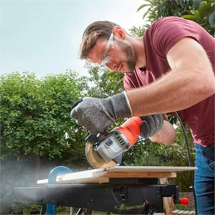 Black+Decker Amoladora angular eléctrica BEG010 - 710 W, Diámetro disco: 115 mm, 12.000 r.p.m. - Un hombre utiliza una amoladora angular con gafas de protección y guantes para cortar una madera.