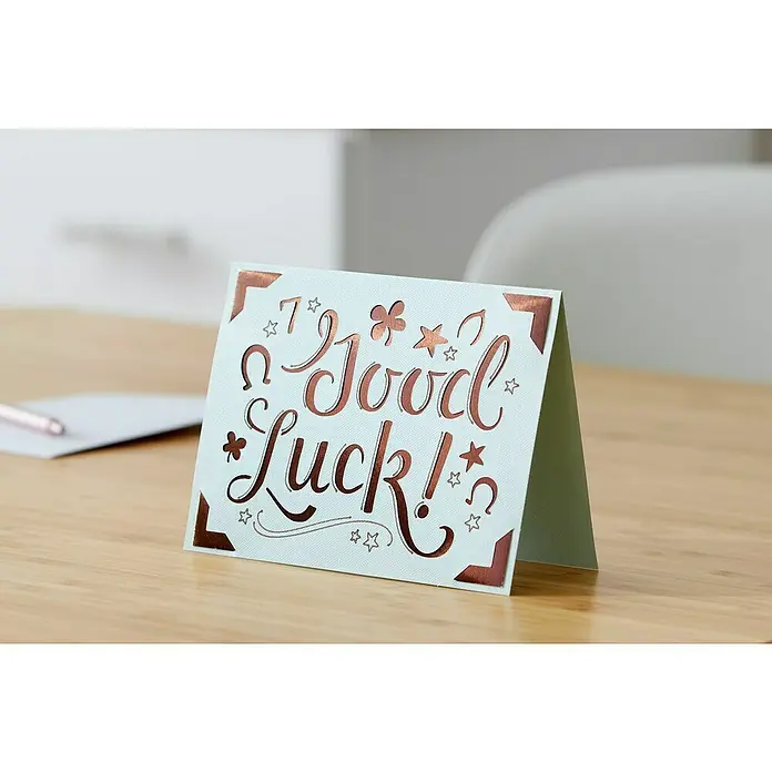 Cricut Joy Schneideplotter Starter Bundle - Bluetooth, Schnittbreite: 10,4 cm, Schnittlänge: 1,2 m - Nahaufnahme einer "Viel Glück!"-Grußkarte mit Glückssymbolen wie Hufeisen, Sternen und Kleeblättern in Kupferfolie auf einem Holztisch.