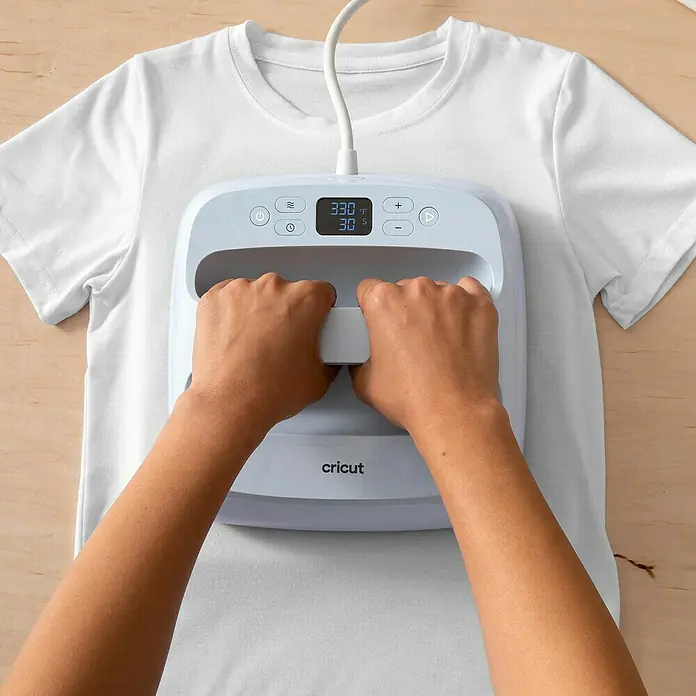 Cricut Joy Xtra Aufbügelfolie Smart Iron-On - Rot, 1 Stk., L x B: 61 x 24,1 cm - Nahaufnahme eines Cricut EasyPress Mini-Bügeleisens auf einem weißen T-Shirt, bedient von zwei Händen.