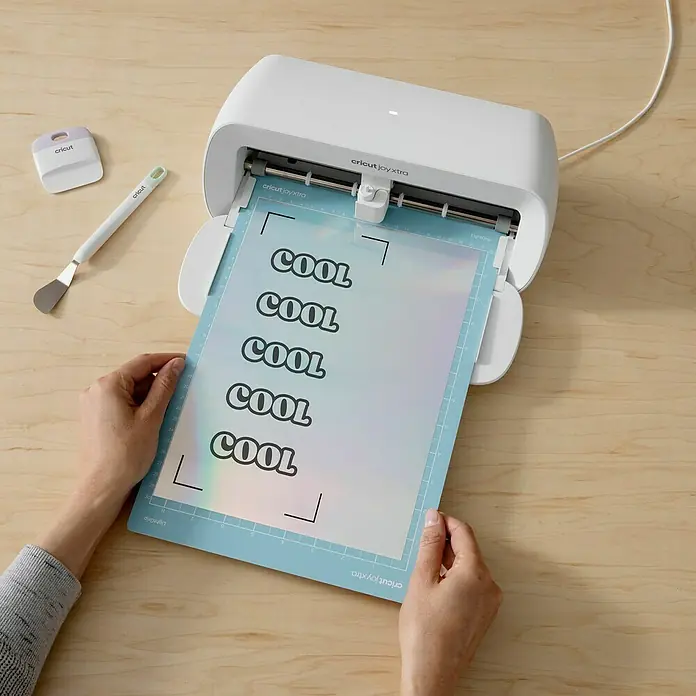 Cricut Vinylfolie Bedruckbar - 10 Stk., Silber, 21 x 29,7 cm - Nahaufnahme eines Cricut Joy Xtra Schneideplotters mit "COOL"-Schriftzug auf einer Schneidematte, ideal für DIY-Projekte.