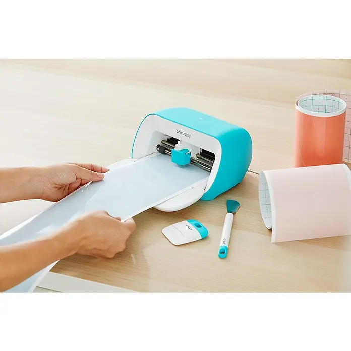 Cricut Joy Schneideplotter Starter Bundle - Bluetooth, Schnittbreite: 10,4 cm, Schnittlänge: 1,2 m - Ein türkisfarbener Cricut Joy Schneideplotter wird mit einer Folie bedient, Werkzeuge und Folienrollen liegen bereit.