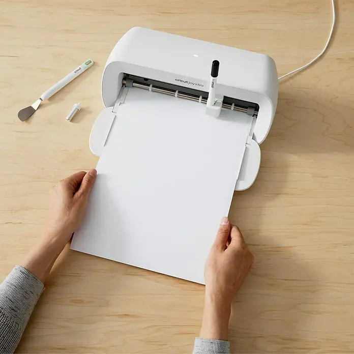 Cricut Joy Xtra Vinylfolie Smart Label Beschreibbar Permanent - 3 Stk., Weiß - Nahaufnahme eines Cricut Joy Xtra Schneideplotters mit einer Person, die ein Blatt Papier hineinlegt, zusammen mit Werkzeugen auf einem Holztisch.