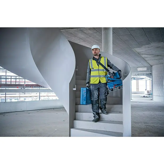 Bosch Professional AMPShare 18V Akku-Nass-Trockensauger GAS 18V-12 MC - Ohne Akku, 18 V, Behältervolumen: 12 l - Ein Bauarbeiter mit gelber Sicherheitsweste und Helm trägt eine Bosch-Werkzeugbox und einen Staubsauger auf einer Wendeltreppe.