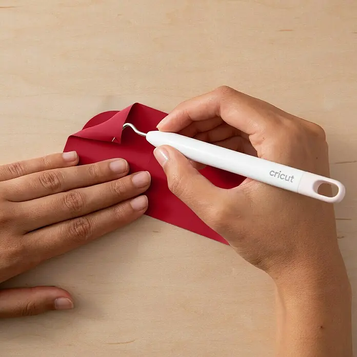 Cricut Joy Xtra Aufbügelfolie Smart Iron-On - Rot, 1 Stk., L x B: 61 x 24,1 cm - Nahaufnahme von Händen, die mit einem Cricut Werkzeug rotes Vinyl bearbeiten, um präzise Details zu entfernen. Perfekt für DIY-Projekte und Bastelarbeiten.